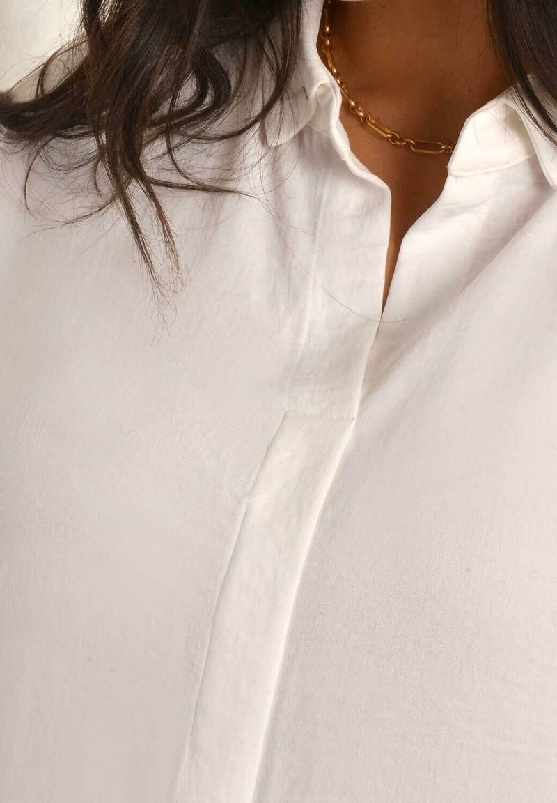 White button-up shirt featuring a collared neckline, soft fabric texture, and a subtle placket detail at the front. Gold chain necklace accent.