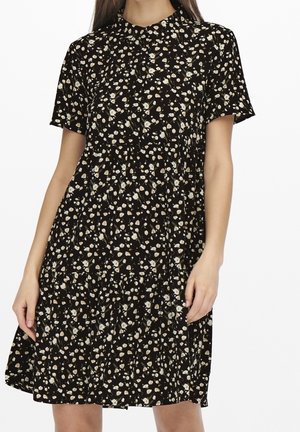 Sort blomsterkjole med korte ærmer, knappelukning ved kraven, lagdelt design, med små hvide og beige blomstermønstre på en mørk baggrund.