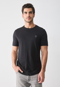 Black short-sleeve t-shirt made from cotton, featuring a round neck and a small emblem on the chest, worn with light grey jeans.
