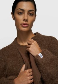 Brauner, flauschiger Mantel mit lockerem Sitz und einem Etikett am Ärmel. Darunter ein eng anliegendes braunes Oberteil mit dezentem Print. Glatte Hautstruktur.