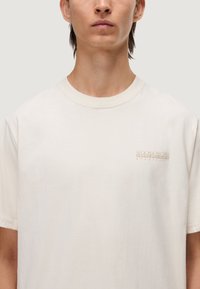 T-shirt a maniche corte di colore crema, realizzato in morbido cotone, con un logo sottile in beige nella parte sinistra del petto. Design classico con scollatura rotonda.
