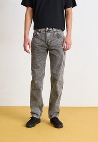 Pár šedých džínů z denimu s vybledlým efektem, rovný střih nohavic, pět kapes a kontrastní šití, nošené s černými botami.