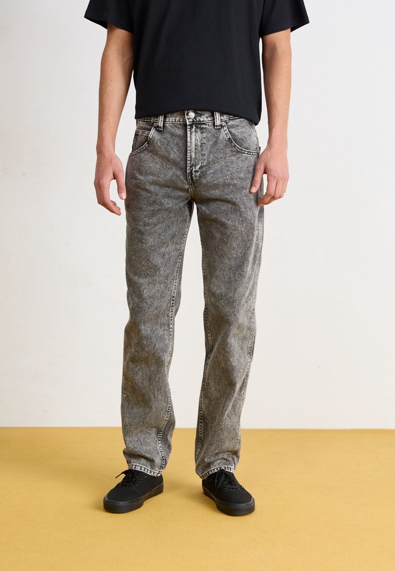 Pár šedých džínů z denimu s vybledlým efektem, rovný střih nohavic, pět kapes a kontrastní šití, nošené s černými botami.