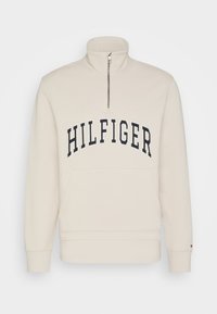 Felpa beige con colletto alto, zip a metà lunghezza e tasca a marsupio frontale. Presenta il logo "HILFIGER" in blu navy sul petto.