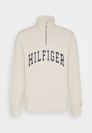 Felpa beige con colletto alto, zip a metà lunghezza e tasca a marsupio frontale. Presenta il logo "HILFIGER" in blu navy sul petto.
