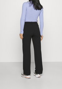 Kobieta z długimi ciemnymi włosami, ubrana w jasnofioletowy sweter z prążkami, czarne szerokie spodnie i srebrne sneakersy, stojąca na tle jednolitego białego tła.