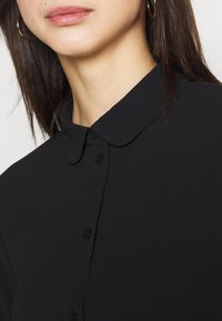 Blusa nera con colletto, realizzata in un tessuto liscio. Presenta una chiusura con bottoni e un design semplice senza motivi o decorazioni.