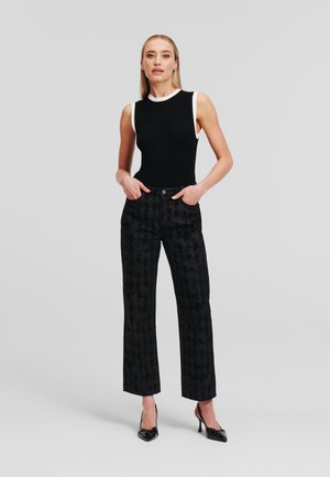 Schwarzes geripptes Tanktop mit weißem Rand, kombiniert mit hochgeschnittenen schwarzen gemusterten Hosen; Model steht mit den Händen in den Taschen und trägt spitze Absätze.