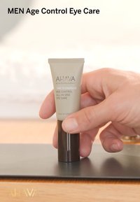 Tube de soin des yeux AHAVA Men Age Control, gris et brun foncé, en forme cylindrique, étiquette indiquant "Il est temps de dynamiser le contrôle de l'âge", capacité de 15 ml.