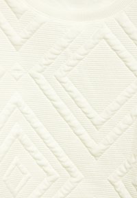 Texturierteter Stoff in einem Off-White-Ton zeigt erhabene Diamant- und Zickzackmuster, die eine haptische Oberfläche mit subtiler Tiefe und Variation schaffen.