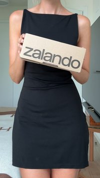 Καφέ χαρτοκιβώτιο με την ένδειξη "zalando" τυπωμένη με μεγάλα γκρι γράμματα· κρατημένο μπροστά από ένα άτομο που φοράει μαύρο άνετο φόρεμα χωρίς μανίκια.