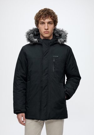 Jeune homme portant un parka d’hiver noir avec une capuche doublée de fourrure et un pantalon beige, debout sur un fond clair uni.