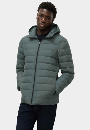 Giacca imbottita di colore verde scuro con chiusura con zip e cappuccio. Presenta una texture trapuntata e tasche laterali. Indossata sopra un top nero.
