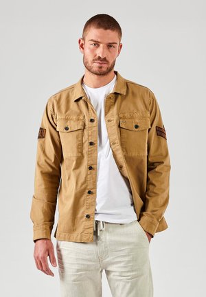 Brun skjorte med knapper og to brystlommer, sorte knapper og et mærke på venstre ærme. Bæres over en hvid t-shirt og lyse bukser.