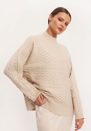 Beżowy sweter w splot kablowy z wysokim kołnierzem, w luźnym fasonie, z prążkowanym wykończeniem i szczegółowymi wzorami. Miękka tekstura i swobodna sylwetka.
