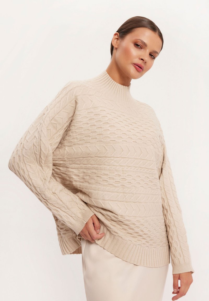 Beżowy sweter w splot kablowy z wysokim kołnierzem, w luźnym fasonie, z prążkowanym wykończeniem i szczegółowymi wzorami. Miękka tekstura i swobodna sylwetka.