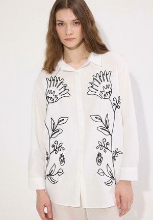 Femme portant une chemise blanche à boutons avec une broderie florale noire sur le devant, debout devant un fond clair uni.