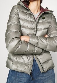 Doudoune vert clair avec fermeture éclair, capuche et doublure bordeaux contrastante. Présente une texture matelassée et des poignets élastiqués.