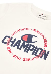 T-shirt in cotone bianco con un logo circolare rosso e blu con il testo "CHAMPION", accompagnato da "AUTHENTIC - ATHLETICWEAR - EST. 1919."