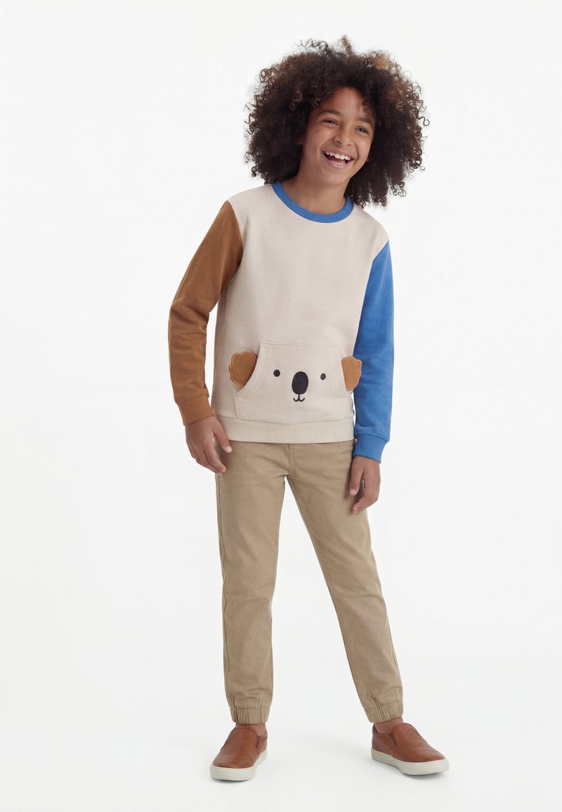 Παιδί με σγουρά μαλλιά που φοράει μπεζ φούτερ με καφέ και μπλε μανίκια και τσέπη με πρόσωπο κοάλα, καφέ παντελόνι και καφέ παπούτσια τύπου slip-on, χαμογελαστό.