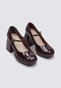 Zapatos Mary Jane de charol burdeos con puntera cuadrada, tacones anchos y detalle de hebilla lateral. Textura suave con un acabado brillante.