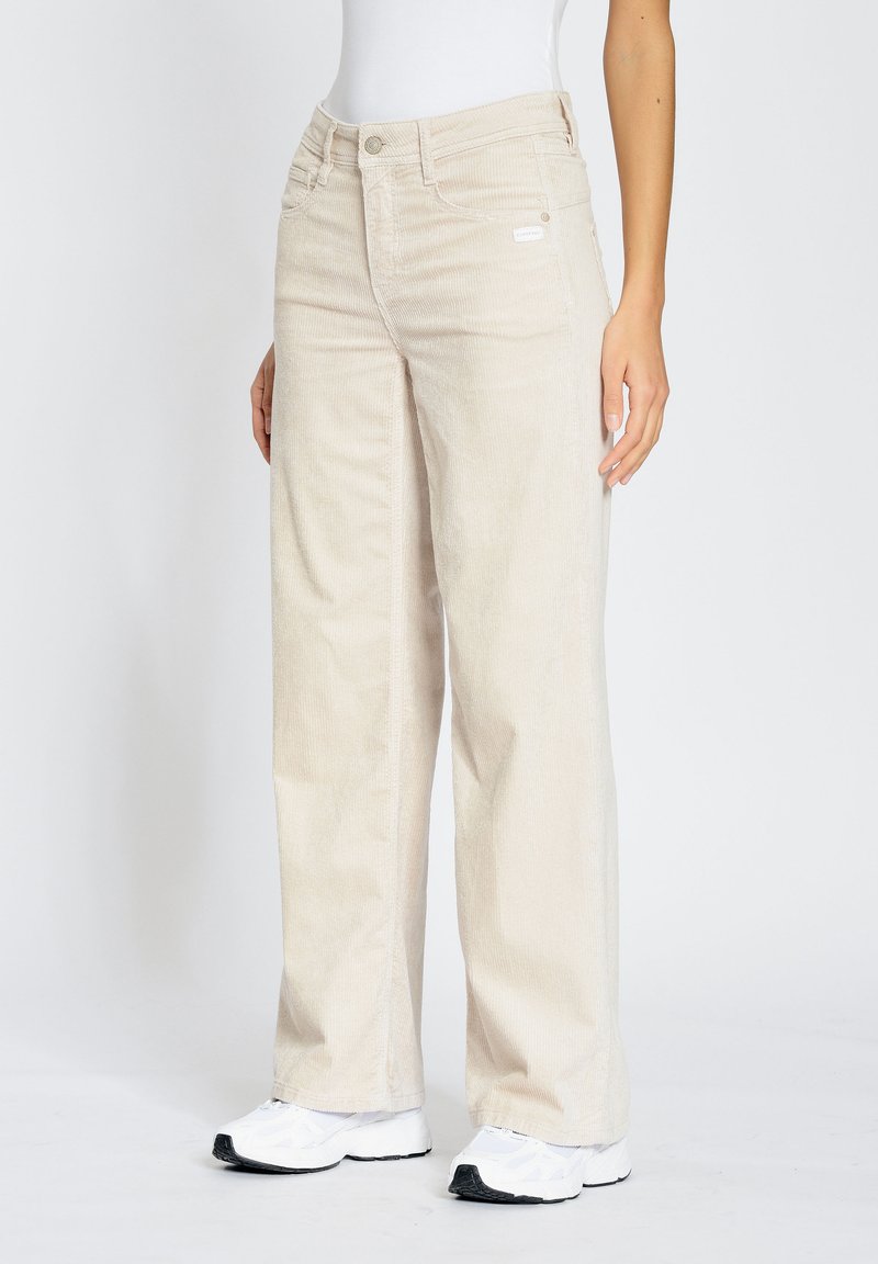 Beige corduroy wide-leg pants with visible textured ribs, featuring a zip fly and front pockets. Worn with white sneakers.