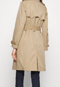 Béžový trenčkot s opaskem, s přezkami na manžetách, zadním dílem a rozparkem, nošený přes tmavé kalhoty, zobrazený zezadu.