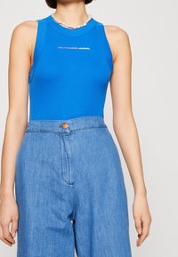 Blå ärmlös bodysuit med rund hals och subtil logotyp. Matchas med ljus tvättad vida denimbyxor med knäppning.