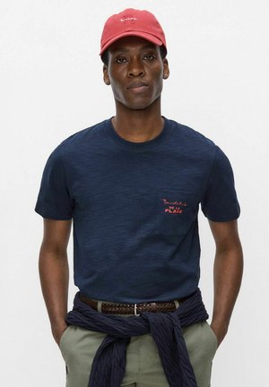 Homme portant une casquette rouge, un t-shirt bleu marine avec texte sur la poche, un pantalon beige et un pull bleu marine noué autour de la taille, debout devant un fond uni.