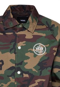 Veste camouflage à col avec boutons-pression, écusson du logo Vans sur la poitrine et doublure intérieure noire visible au niveau du col.