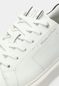 Λευκό δερμάτινο αθλητικό παπούτσι με επίπεδα λευκά κορδόνια, μικρό λογότυπο στη γλώσσα και χρυσή λεπτομέρεια στην τρύπα για τα κορδόνια. Ομαλή υφή με διακριτικές λεπτομέρειες ραφής.