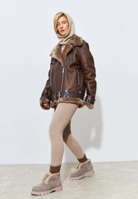 Veste en cuir brun avec doublure en shearling, leggings beiges à côtes et bottes grises avec accents en fausse fourrure. Le mannequin marche sur une surface claire.