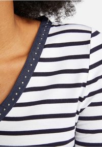 Randig V-ringad skjorta i vitt och marinblått, med metalliska nitar längs halsringningen. Mjuk tygkvalitet med avslappnad passform.