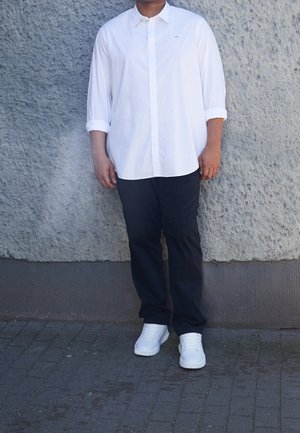 Mand iført en hvid langærmet skjorte med opkrogede manchetter, sorte bukser og hvide sneakers, der står op ad en struktureret grå væg.