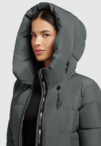 Veste en duvet gris foncé, dotée d'une grande capuche, d'une fermeture éclair et de cordons de serrage élastiques. Texturée avec un motif matelassé et des accents de logo.
