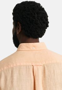 Helles Pfirsich leinen Hemd mit einem Knopfkragen, ausgestattet mit einem Rückenschlitz und feiner Textur. Knöpfe und Nähte sind am Kragen sichtbar.