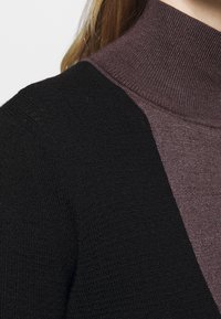 Närbild av en person som bär en svart stickad cardigan över en lila räfflad tröja med hög hals, med blont hår delvis synligt.