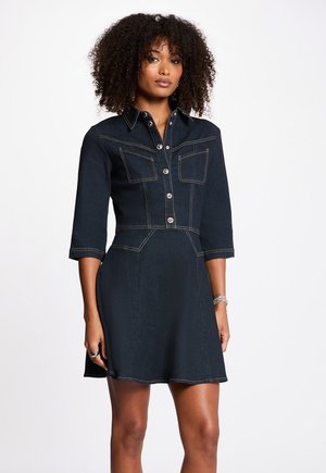 Frau mit lockigem Haar, die ein dunkles Jeanskleid mit Knöpfen, dreiviertel Ärmeln, Fronttaschen und einem ausgestellten Rock trägt, vor weißem Hintergrund stehend.