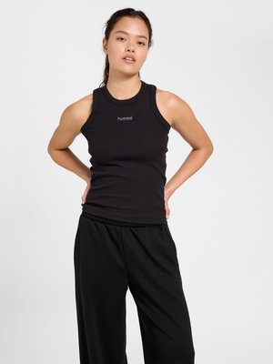 Młoda kobieta stoi z rękami na dolnej części pleców, ubrana w czarny, bezrękawnikowy prążkowany top i luźne czarne spodnie na jednolitym tle.