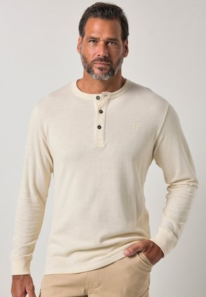 Mand med kort mørkt hår og skæg iført en cremefarvet langærmet henley-shirt og beige bukser, står med den ene hånd i lommen.