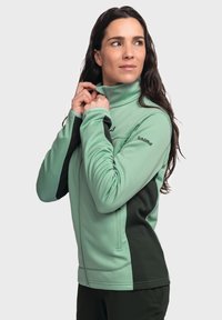 Grüne Fleecejacke mit hohem Kragen, ausgestattet mit schwarzen Seitenpaneelen. Der Stoff hat eine strukturierte Oberfläche und Fronttaschen mit Reißverschluss.