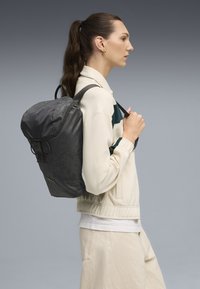Sac à dos gris, imperméable, avec un sommet arrondi, des bretelles réglables et une fermeture à boucle. Présente un motif de logo subtil sur le tissu.