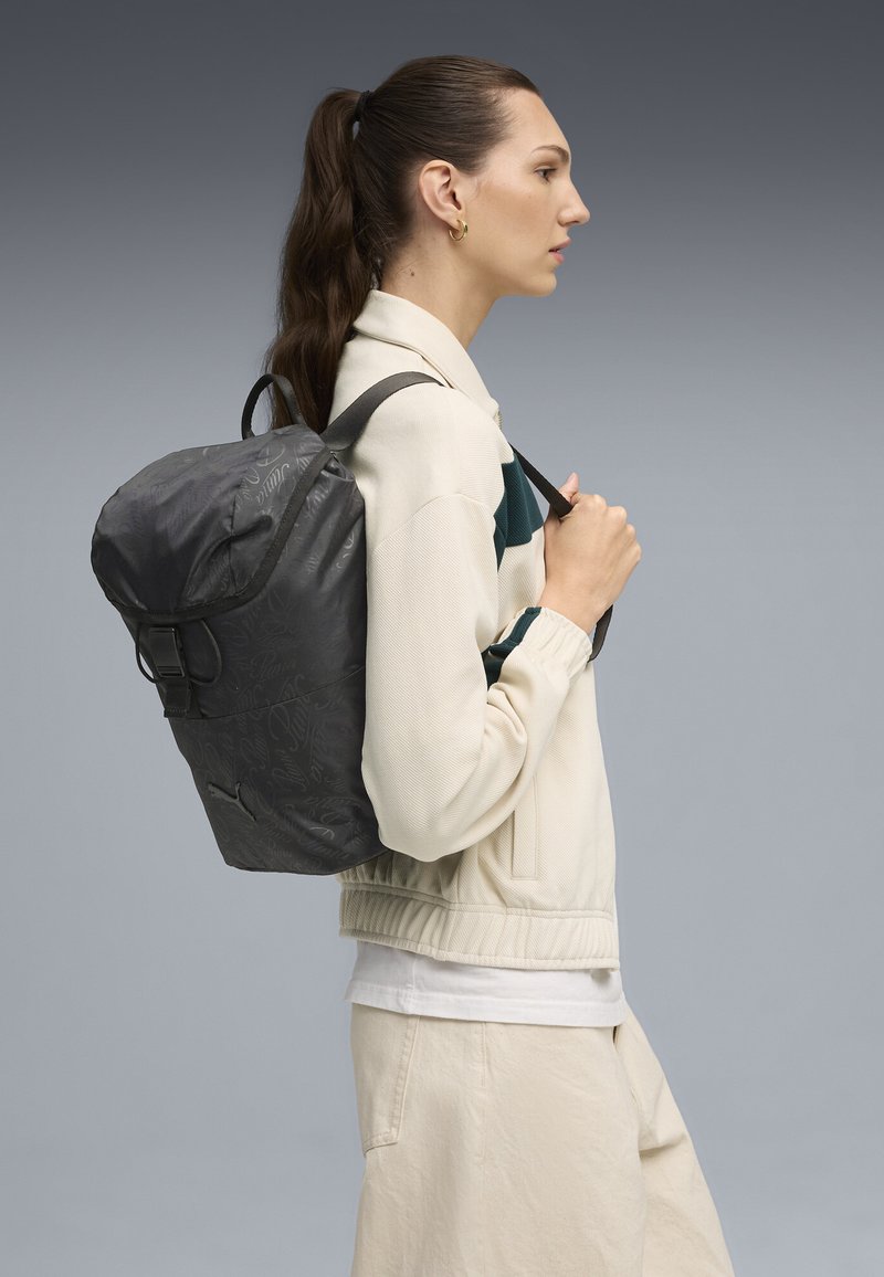 Sac à dos gris, imperméable, avec un sommet arrondi, des bretelles réglables et une fermeture à boucle. Présente un motif de logo subtil sur le tissu.