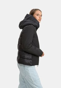 Giacca a vento nera con design imbottito e un grande cappuccio. Presenta una finitura liscia e chiusura con zip, indossata sopra jeans blu chiaro.