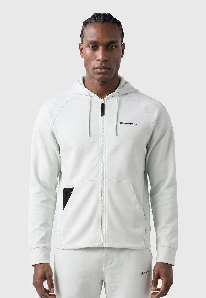 Homme aux cheveux tressés portant un sweat à capuche zippé Champion gris clair et un pantalon assorti, debout devant un fond uni.