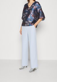 Blumenspitze in marineblau, lila und silbernen Tönen; weite, hellblaue Hosen; silberne, spitz zulaufende Ankle Boots.