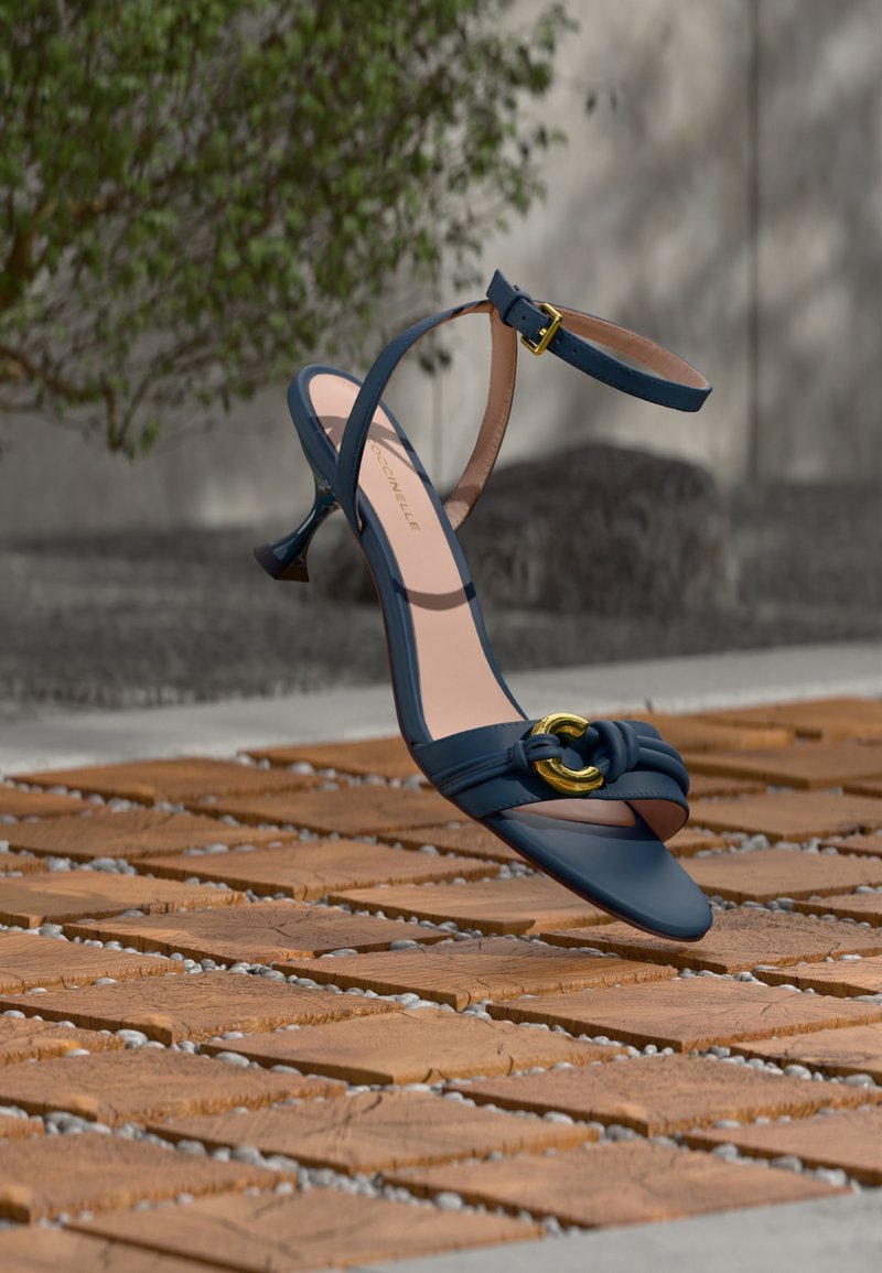 Sandale à talon bleu marine avec bride à la cheville et détail boucle dorée flottant au-dessus d'un sol en bois et motif en pierre en plein air.