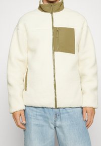 Man draagt een crèmekleurig fleecejack met een lichtbruin ritsvak op de borst en een kraag, gecombineerd met lichtblauwe spijkerbroek.