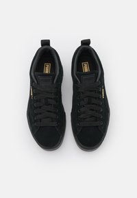 Schwarze Wildleder-Sneaker mit flachen schwarzen Schnürsenkeln, metallisch goldfarbenem Branding, gepolstertem Schaft und strukturiertem Gummisohle. Keine sichtbaren Muster oder Akzente.