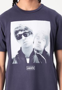 Navy blue cotton t-shirt featuring a graphic print of two figures in black and white with the text "oasis" in a rectangular box at the bottom.